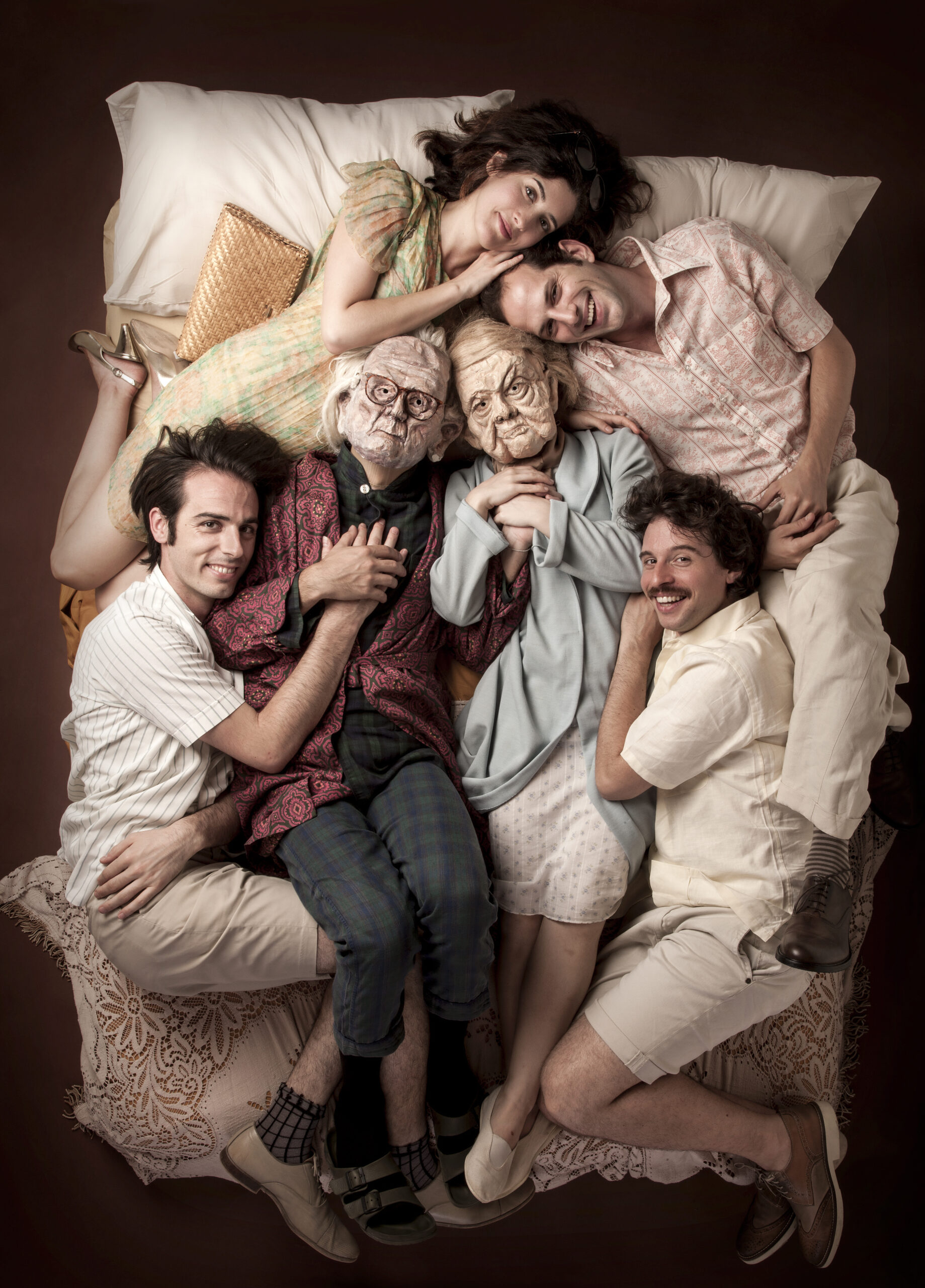 Sei attori si abbracciano su un letto, due con maschere da anziani.