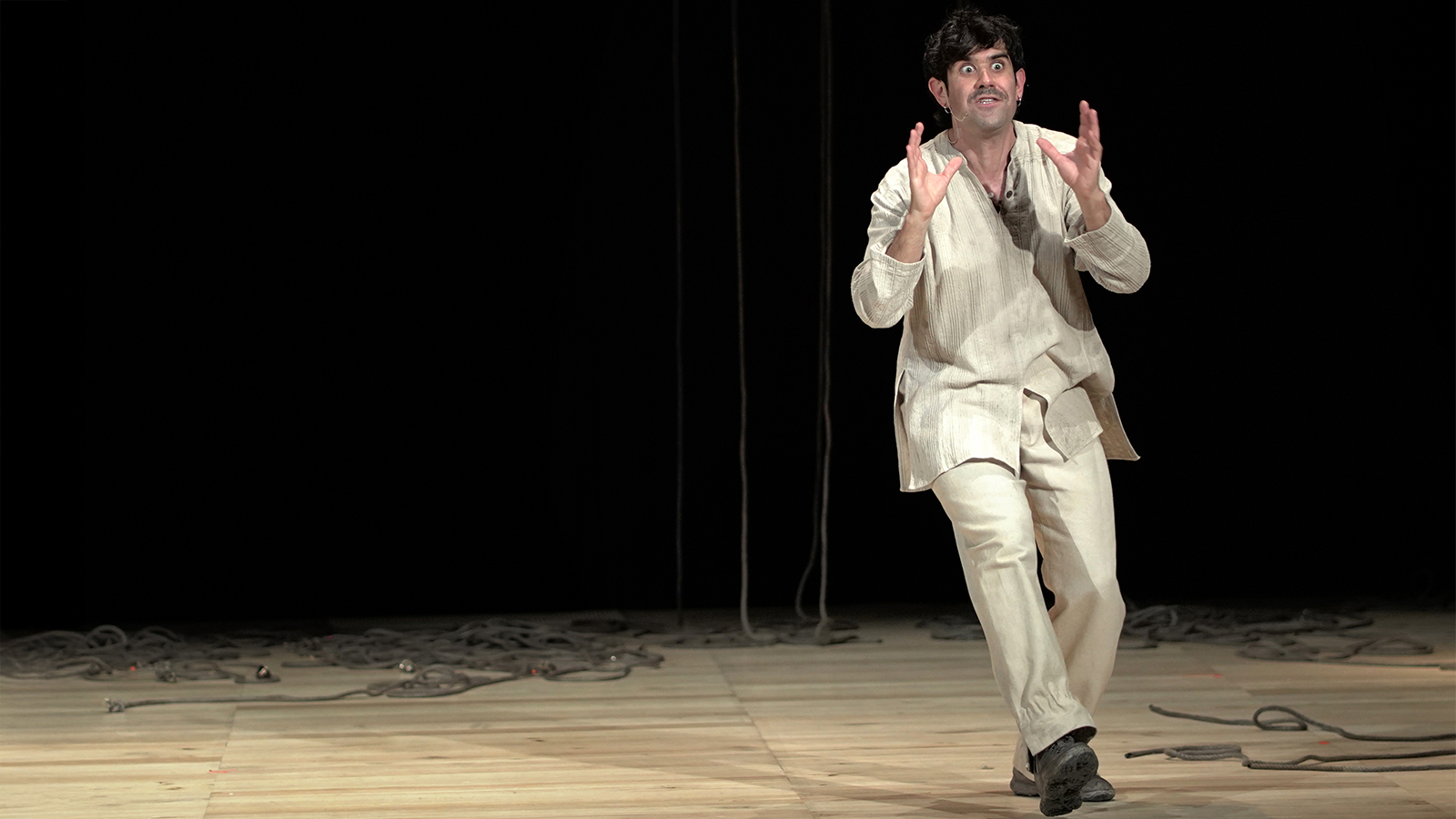 Matthias Martelli, in abiti chiari di tela, avanza verso il pubblico con occhi sgranati e mani aperte, su un palco cosparso di corde.